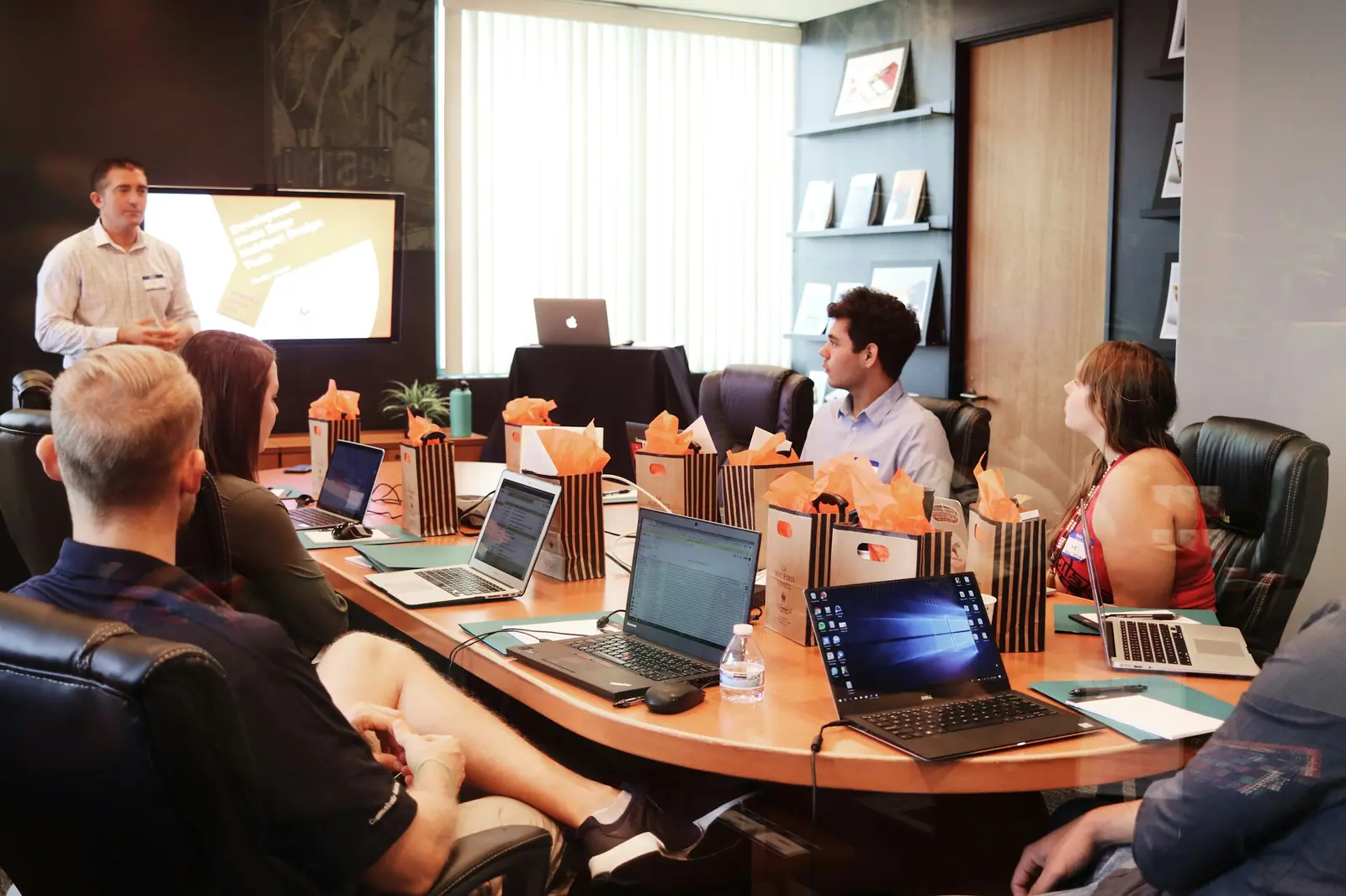 Reunión de trabajo en una sala con una mesa curva: una persona presenta mientras el equipo escucha con sus laptops